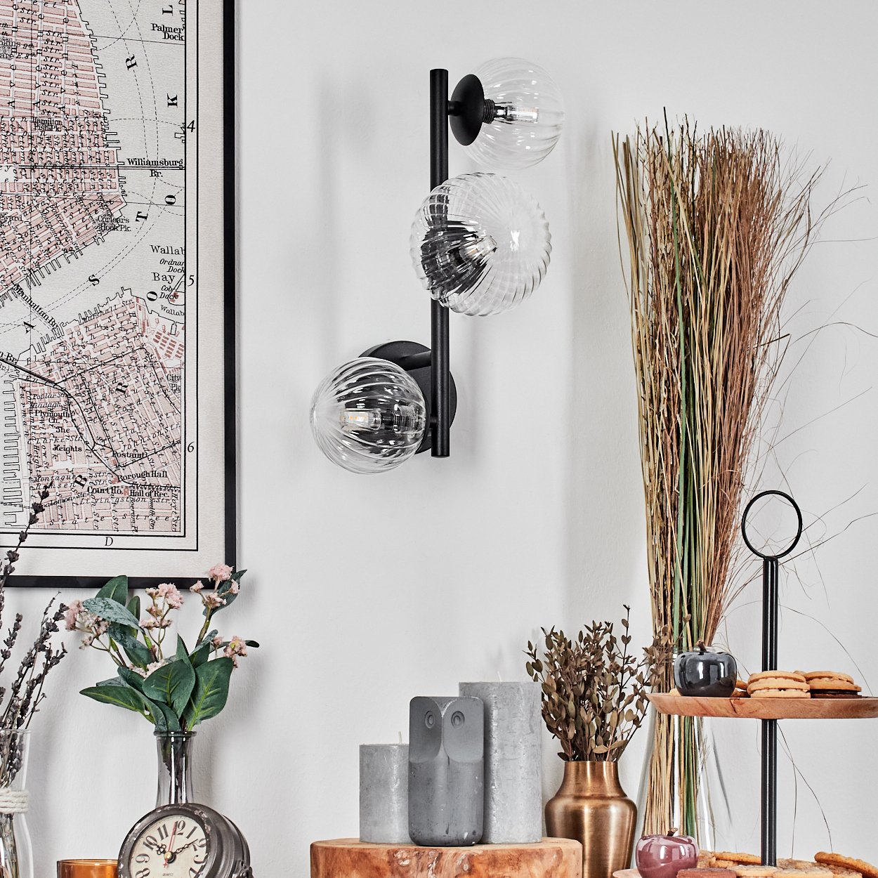 Eine moderne Wandlampe mit drei Glaskugeln. Eine Karte, Blumen, Kerzen und Dekorationsartikel sind auf einer Holzoberfläche angeordnet.