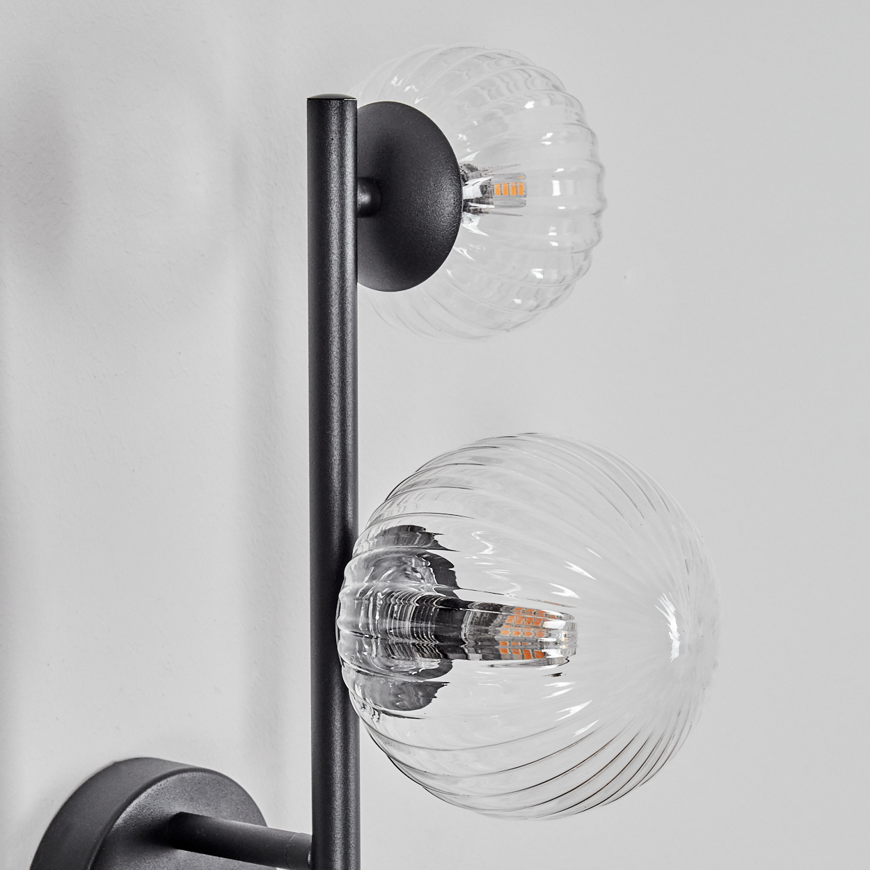 Eine moderne Wandleuchte mit zwei transparenten, gerippten Glaskugelschirmen. Die Halterung ist schwarz und an einer weißen Wand befestigt.