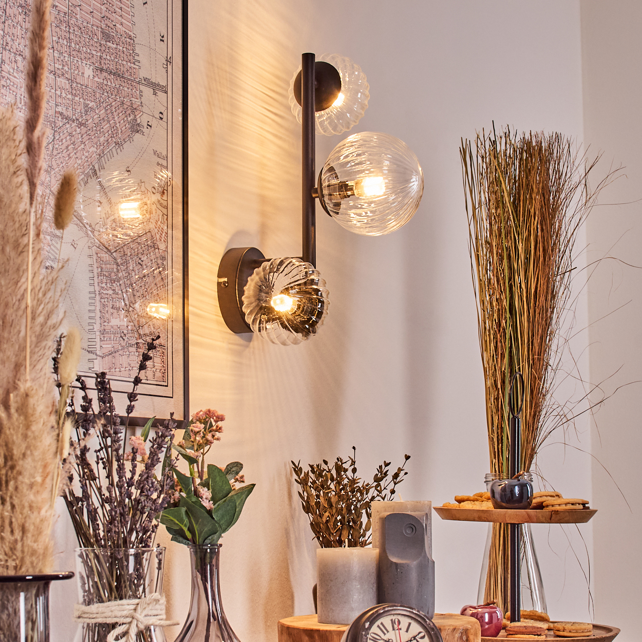 Eine dekorative Wandlampe mit drei Glaskugeln an einer weißen Wand. Vasen mit Blumen und dekorative Gegenstände im Vordergrund.