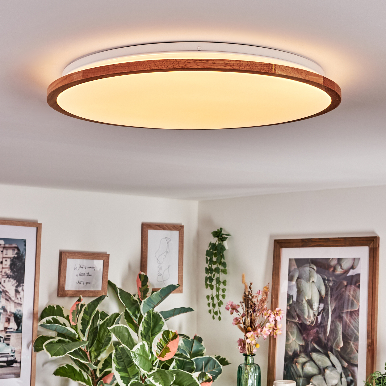 Eine runde Deckenlampe mit Holzrahmen leuchtet. Darunter befinden sich gerahmte Bilder, Pflanzen und eine Blumenvase.