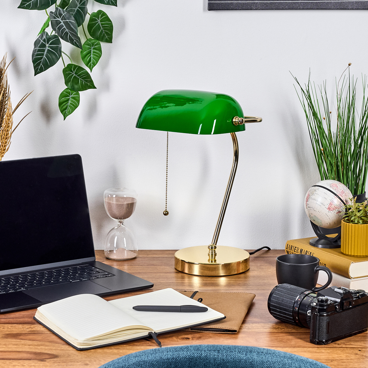 Eine Schreibtischszene mit grüner Lampe, Laptop, Notizbuch, Kamera und Globus. Grüne Pflanzen hängen von oben. Eine schwarze Kaffeetasse ist sichtbar.