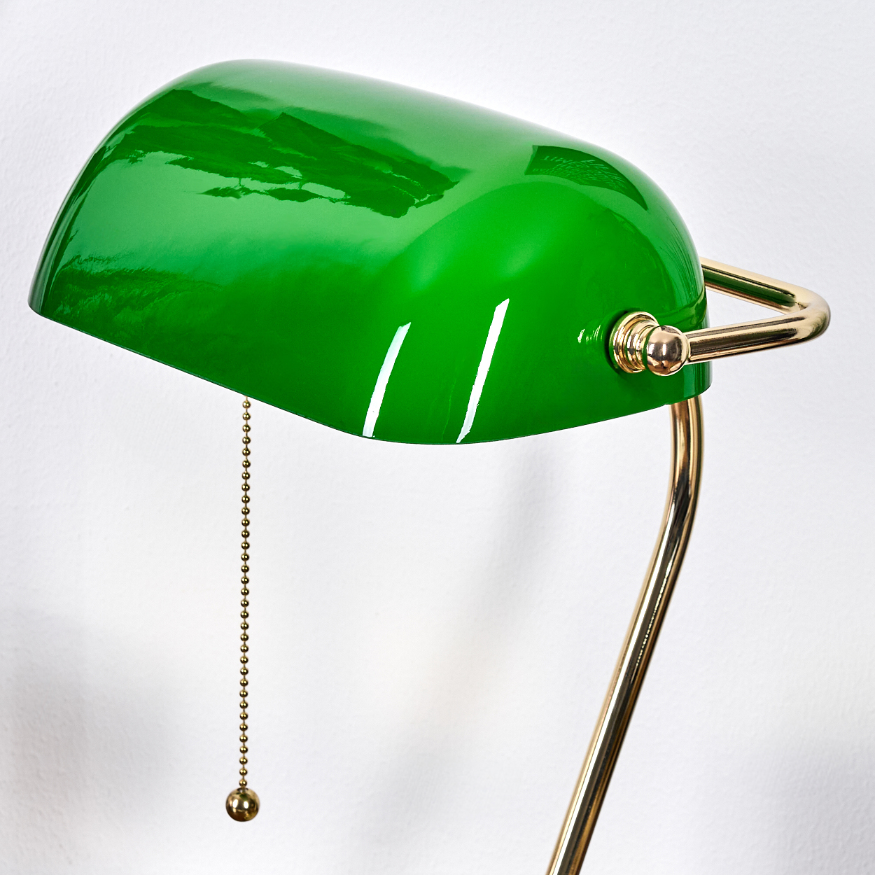 Eine grüne Bankerlampe mit goldenem Fuß und Akzenten hängt vor einer weißen Wand. Eine Kette mit Kugel hängt von der Lampe.