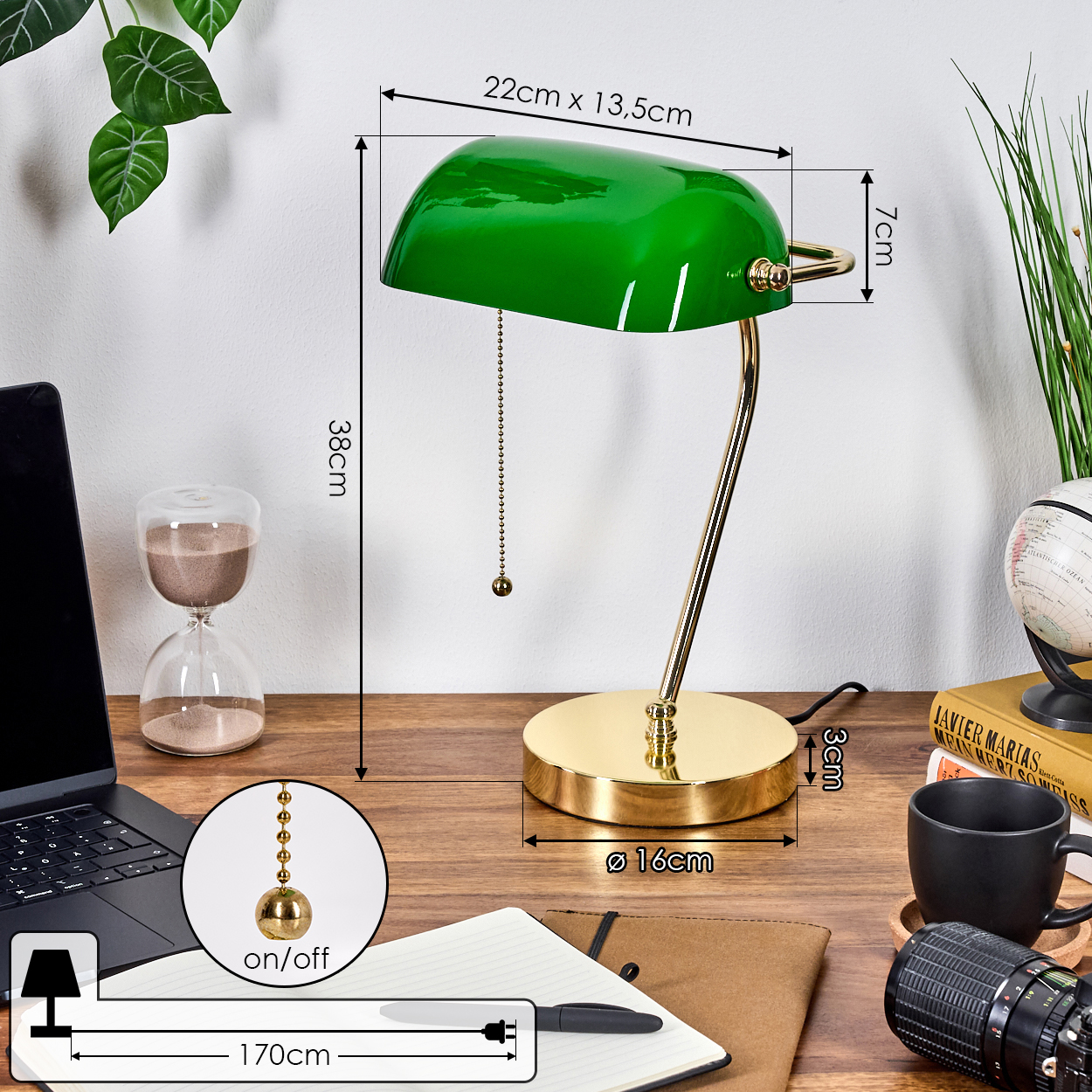 Eine grüne Bankerlampe mit goldenen Akzenten steht auf einem Holztisch. Ein Laptop, eine Sanduhr, eine Kaffeetasse und eine Kamera sind ebenfalls zu sehen.