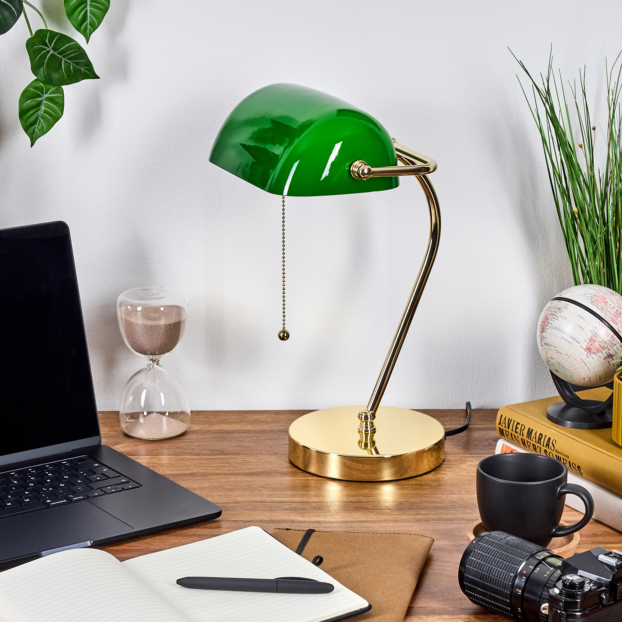 Ein Schreibtisch mit grüner Lampe, Laptop, Sanduhr, Globus, Büchern, Tasse, Kamera und Notizbuch.