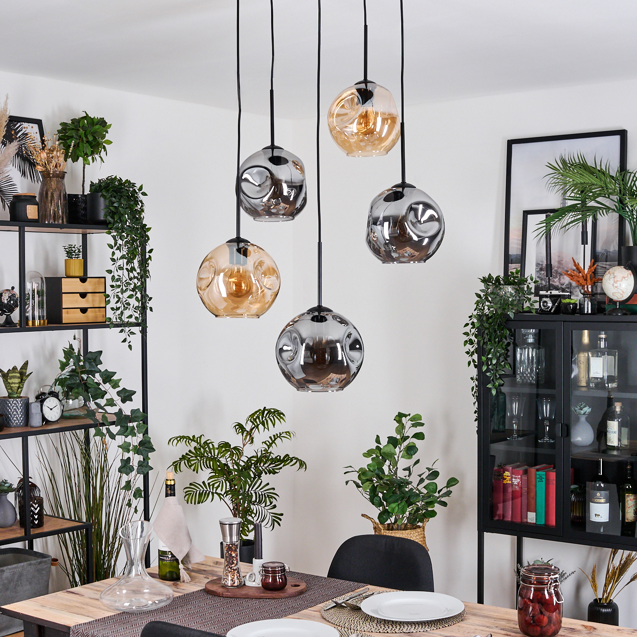 Esszimmer mit Holztisch, Hängelampen, Pflanzen und einem schwarzen Schrank mit Büchern und Dekorationen.
