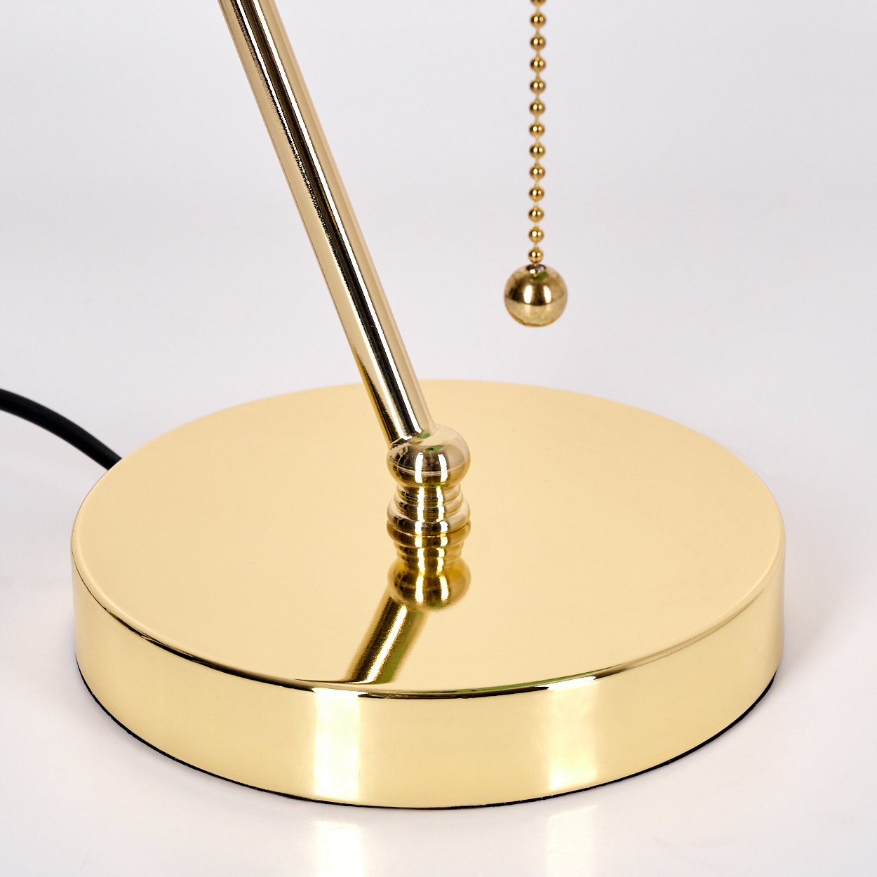 Nahaufnahme eines goldenen Lampenfußes. Eine goldene Stange erstreckt sich nach oben. Eine Kette mit Goldkugel hängt herunter. Schwarzes Netzkabel.