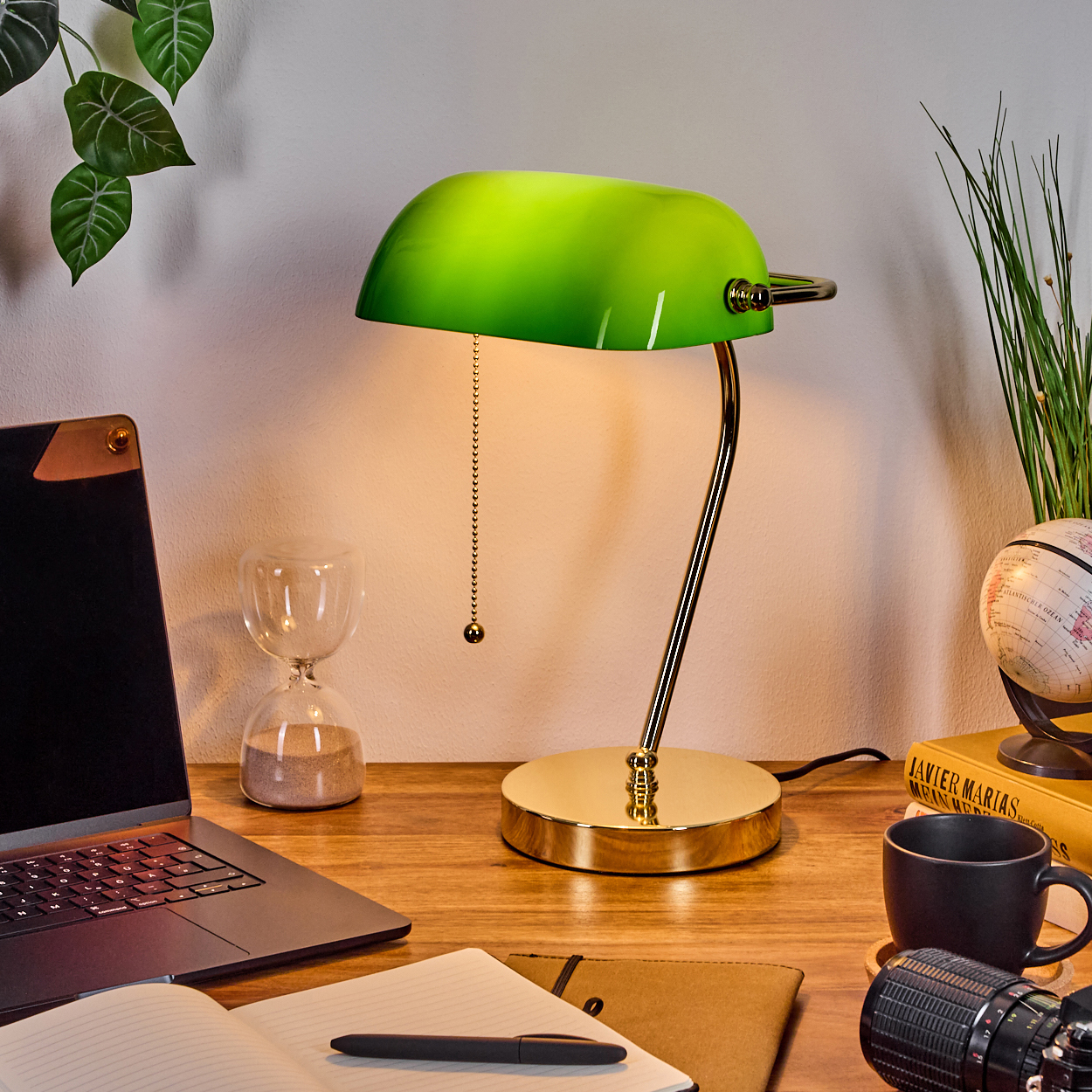 Eine grüne Banker-Lampe steht auf einem Holztisch mit Laptop, Notizbuch, Sanduhr, Kaffeetasse und Globus.