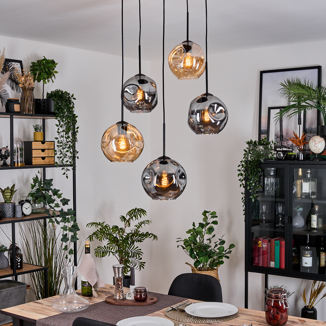 Esszimmer mit hängenden Kugelleuchten. Tisch mit Pflanzen und Dekor. Regale und Schrank sind sichtbar, gefüllt mit Gegenständen.