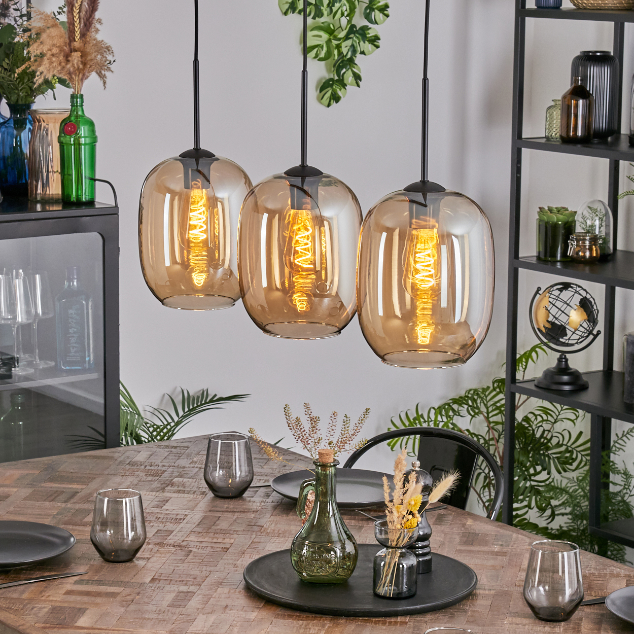 Drei Pendelleuchten mit bernsteinfarbenen Glasschirmen hängen über einem Holztisch, der mit Tellern, Gläsern und einem Mittelstück gedeckt ist.