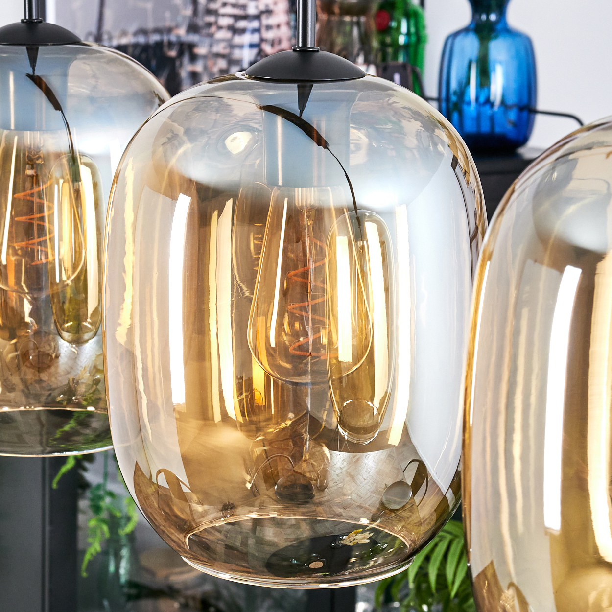 Nahaufnahme von drei Glas-Pendelleuchten mit warm getönten Glühbirnen. Im Hintergrund Vasen und Grün, mit Stadtbild-Reflexion.