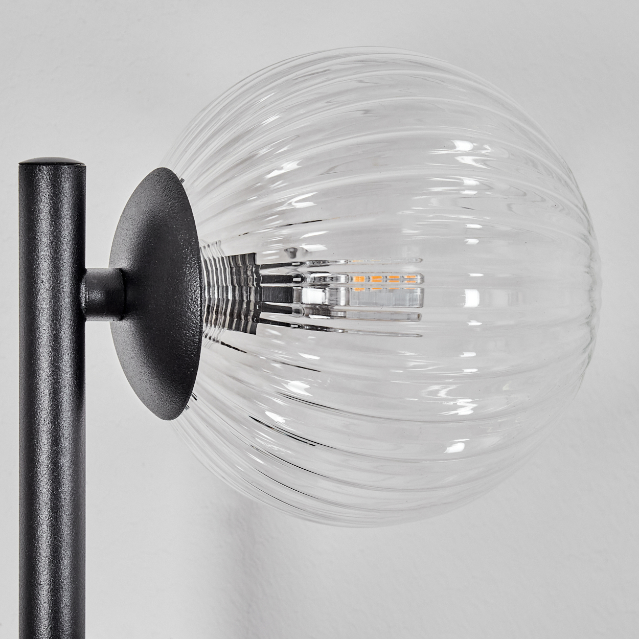 Nahaufnahme einer Lampe. Sie hat eine gerippte Glaskugel und einen schwarzen Metallständer. Der Hintergrund ist eine weiße Wand.