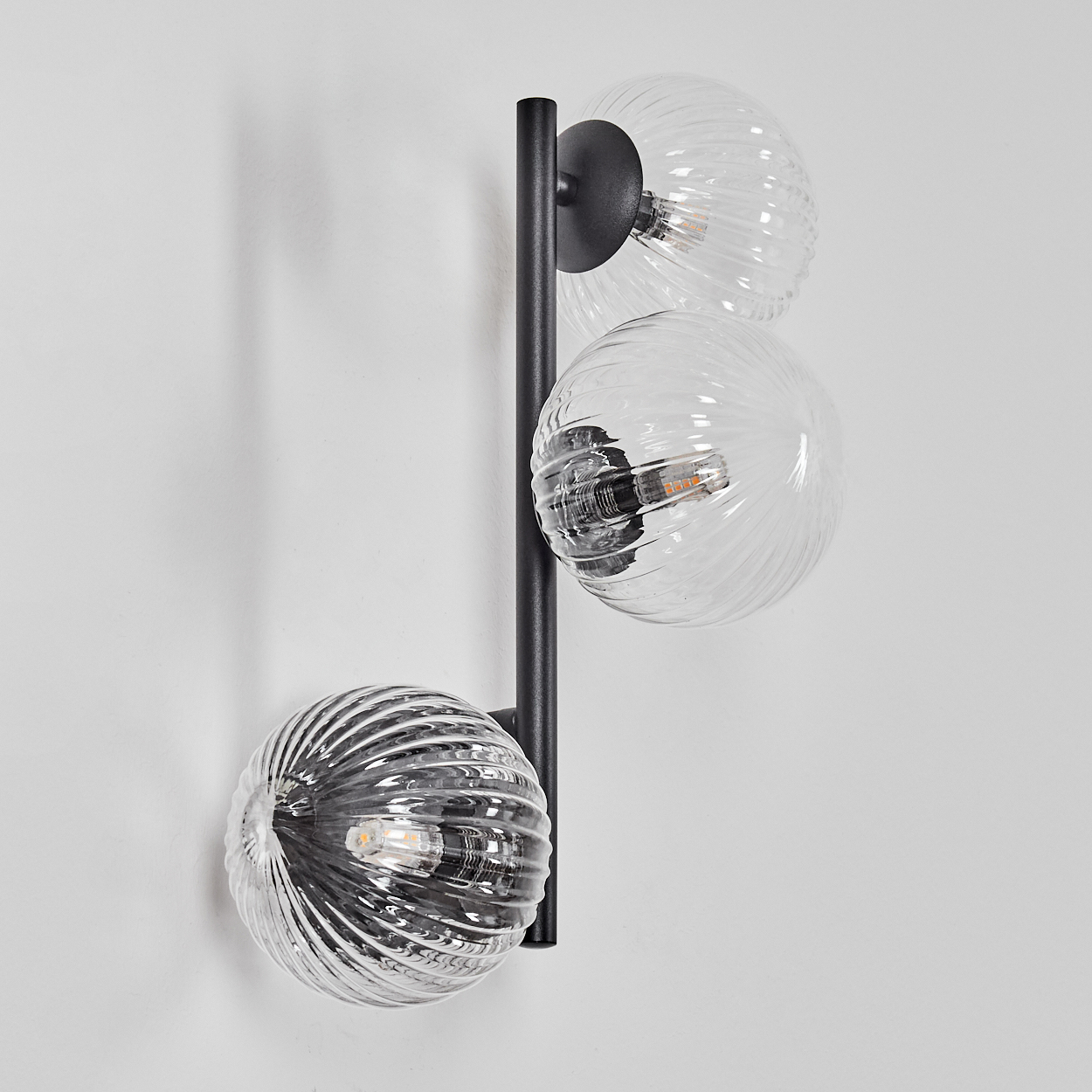 Eine moderne Wandlampe mit drei Glaskugeln. Die Lampe ist an einem schwarzen Metallrahmen an einer weißen Wand befestigt.