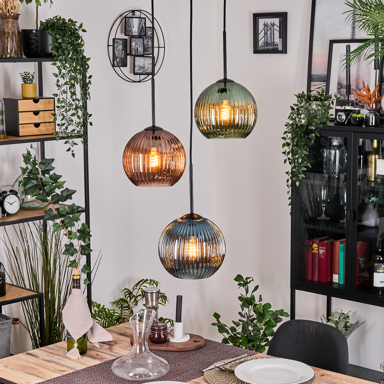 Esszimmer mit drei Hängelampen. Tisch mit Dekanter, Teller und Besteck. Pflanzen und ein Schrank im Hintergrund.