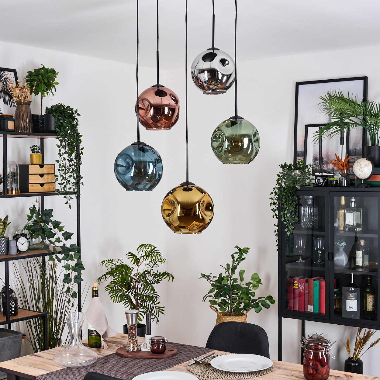 Esszimmer mit fünf bunten Pendelleuchten. Pflanzen und Dekorationen umgeben einen Holztisch, der für eine Mahlzeit gedeckt ist.