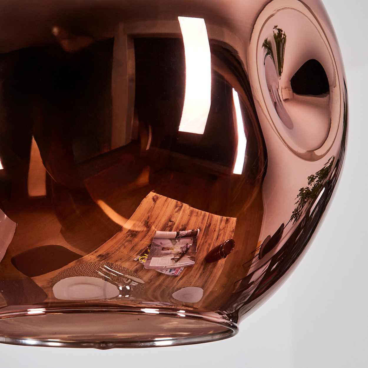 Eine kupferfarbene, reflektierende Leuchte ist aufgehängt. Sie reflektiert einen Holztisch mit einer Zeitschrift, einem Teller und einer Gabel.
