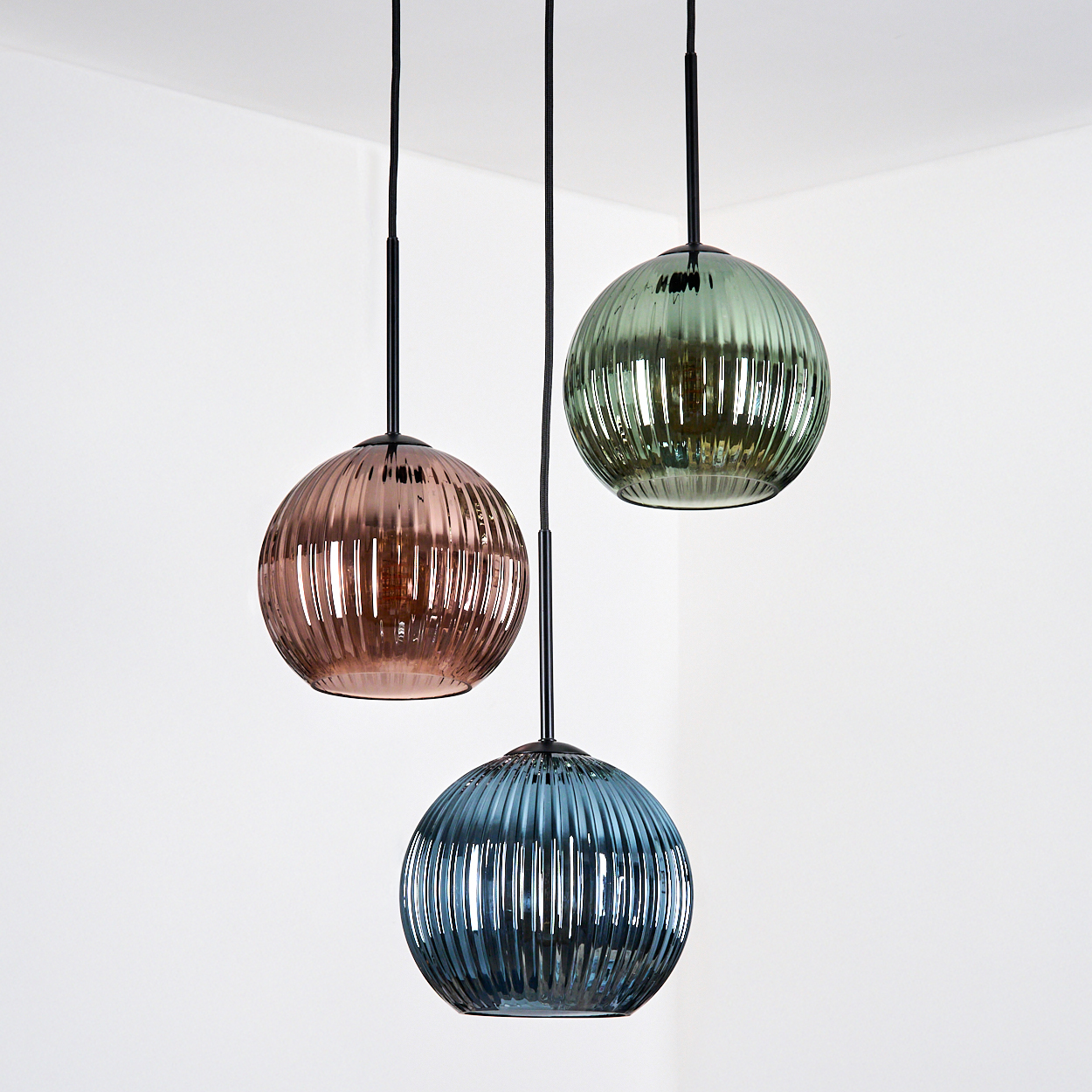 Drei gläserne Pendelleuchten in verschiedenen Farben: Rosa, Grün und Blau. Sie hängen vor einer weißen Wand.