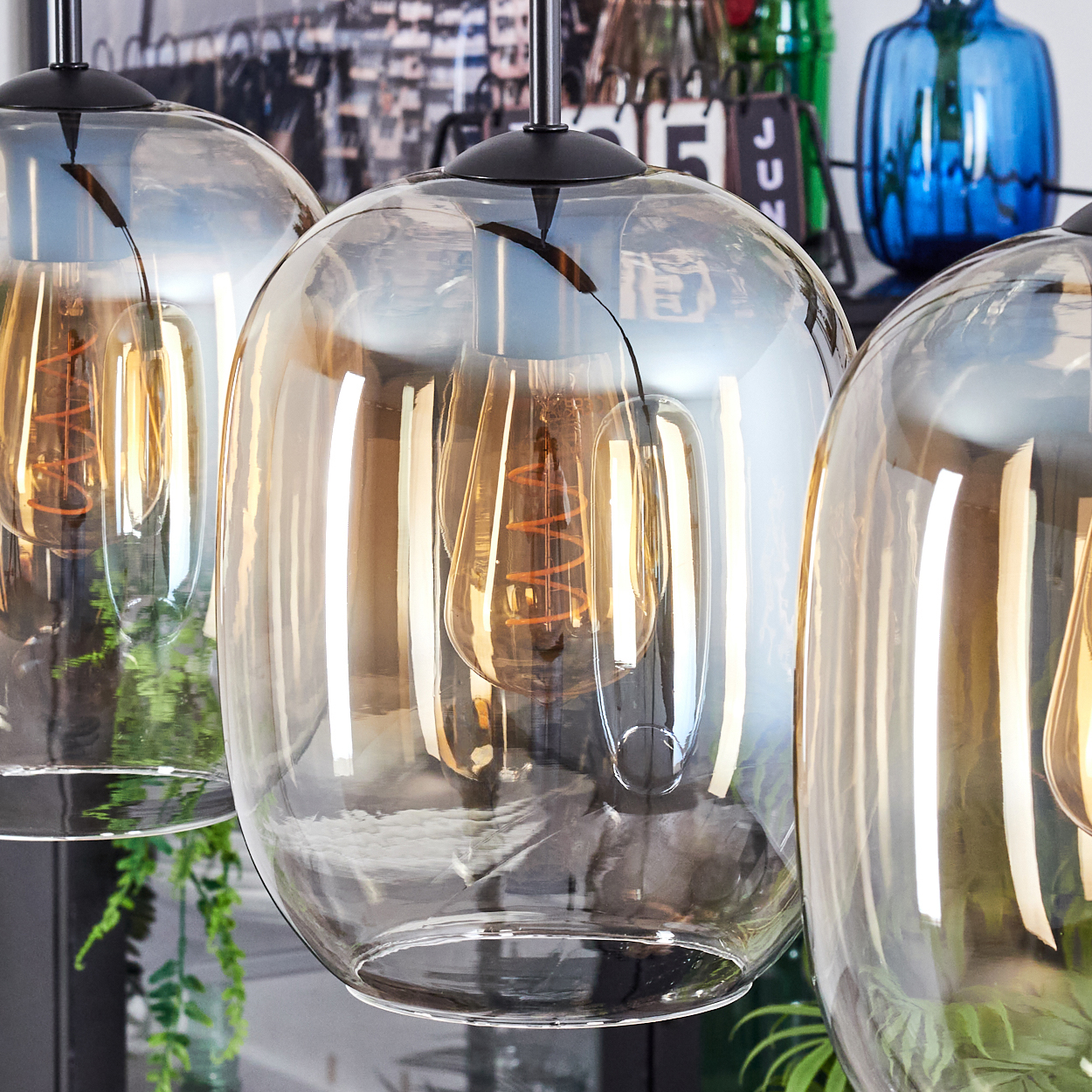 Drei Glas-Pendelleuchten mit warmen Glühbirnen. Im Hintergrund verschwommenes Dekor und Pflanzen. Die Schirme sind klar und kugelförmig.