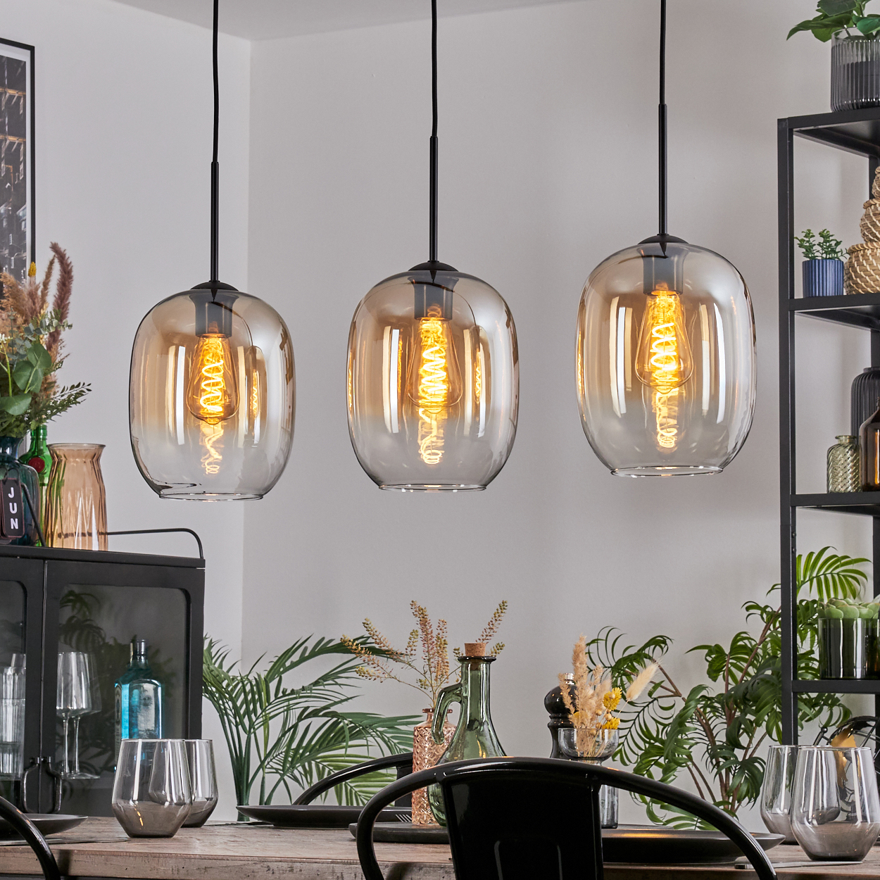 Drei Pendelleuchten mit bernsteinfarbenen Glasschirmen hängen über einem Tisch, der für das Essen gedeckt ist, umgeben von Pflanzen und Dekorationen.