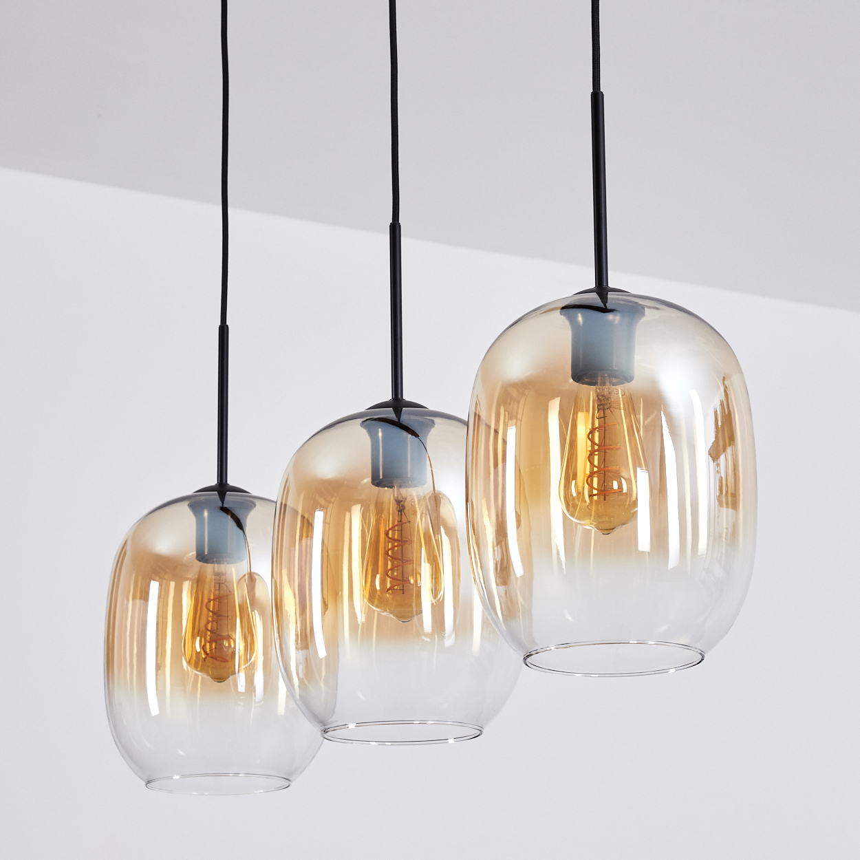 Drei Pendelleuchten mit bernsteinfarbenen Glasschirmen und Edison-Glühbirnen hängen vor weißem Hintergrund von der Decke.