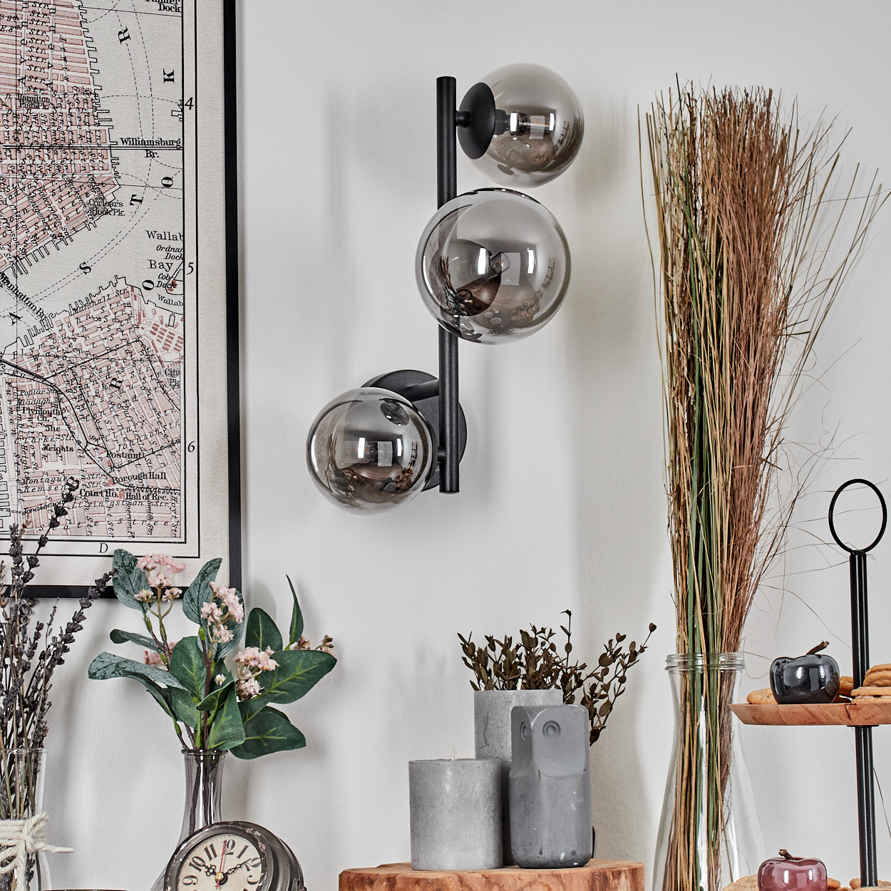 Eine Wand mit einer Karte, einer schwarzen Lampe mit drei Glaskugeln, Blumen, Kerzen und dekorativen Gegenständen. Der Hintergrund ist weiß.