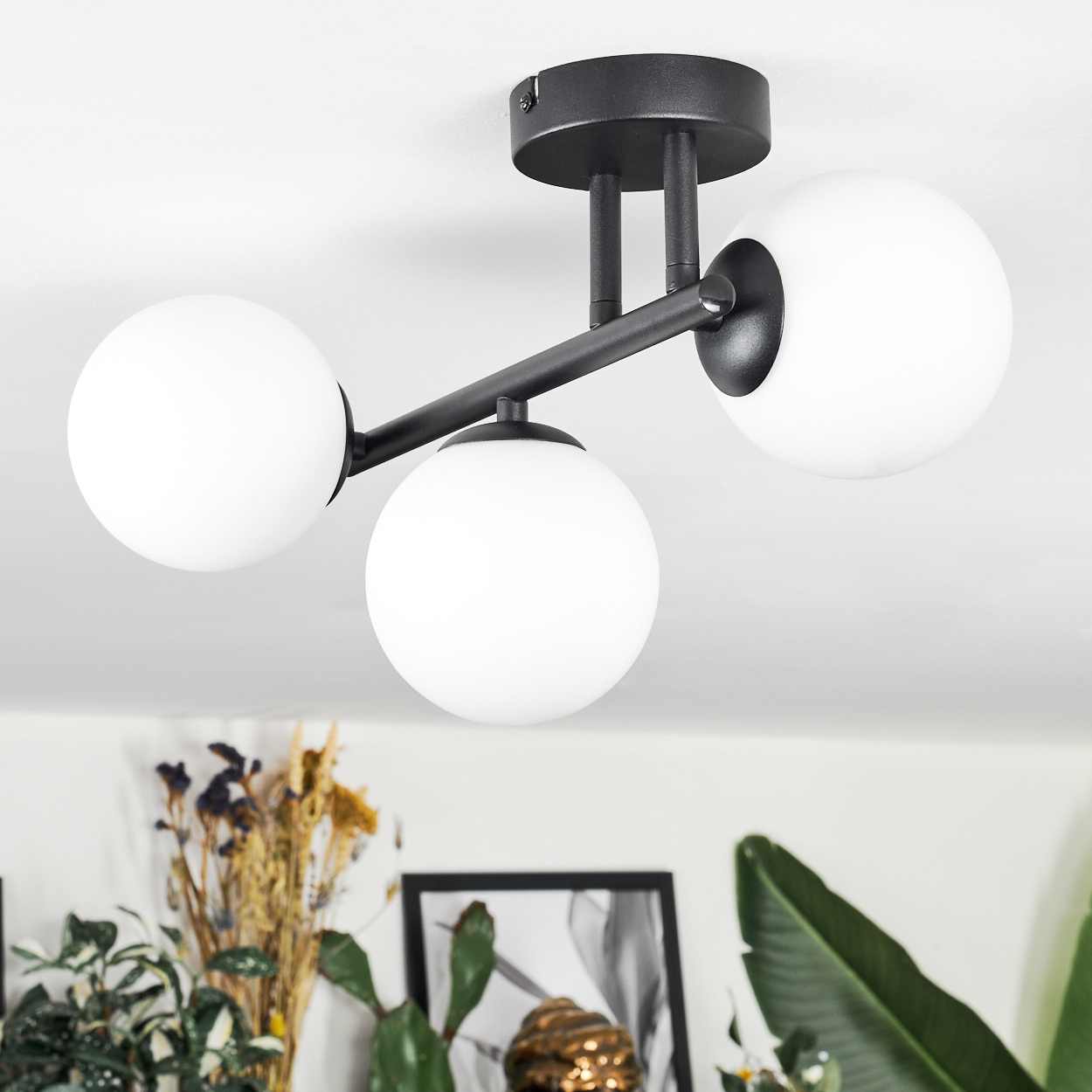 Eine moderne Deckenlampe mit drei weißen Kugeln und einem schwarzen Rahmen. Der Hintergrund zeigt Pflanzen und ein gerahmtes Bild.