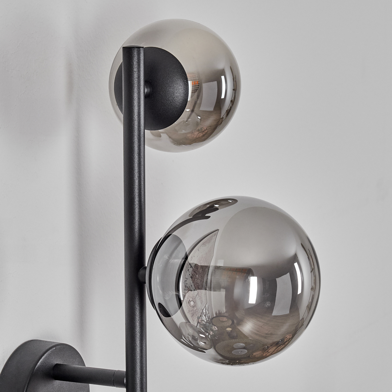 Eine moderne Wandlampe mit zwei rauchigen Glaskugeln. Ein schwarzer Metallrahmen trägt die Kugeln vor einer weißen Wand. Reflexionen sind sichtbar.