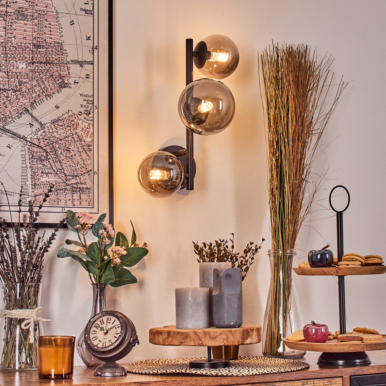 Eine Zimmerecke mit dekorativen Gegenständen: eine Karte, eine Wandlampe, Vasen mit Blumen und Gras, Kerzen, eine analoge Uhr und ein zweistufiges Tablett.