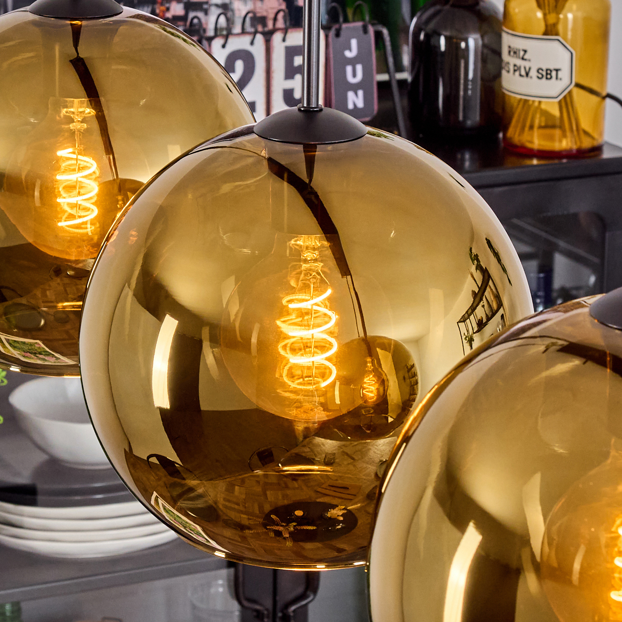 Nahaufnahme von drei kugelförmigen Lampen aus bernsteinfarbenem Glas mit Wendelglühbirnen. Hintergrund: Kalender, Flaschen und Teller.
