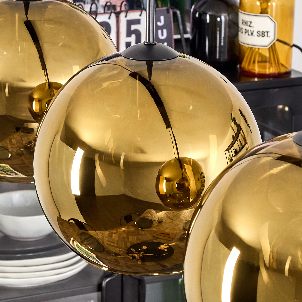 Drei goldene, kugelförmige Pendelleuchten hängen. In dem Glas sind Reflexionen zu sehen. Andere Objekte sind im Hintergrund teilweise sichtbar.