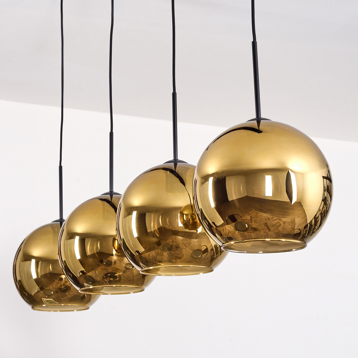 Vier goldene, kugelförmige Pendelleuchten, die an schwarzen Schnüren von einer weißen Decke hängen. Reflektierende Oberflächen.