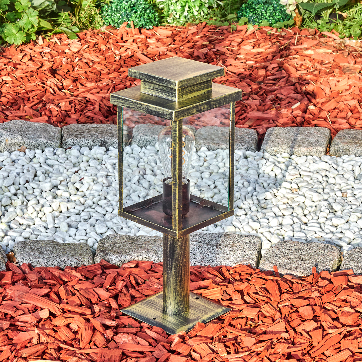Eine dekorative Außenlampe mit Glasgehäuse steht auf roter Mulch, weißen Steinen und Granitkanten. Im Hintergrund grüne Pflanzen.
