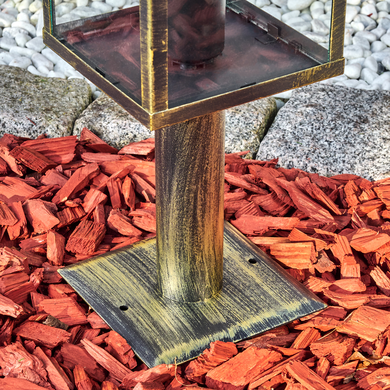 Eine Metalllaterne mit quadratischem Glasaufsatz, auf einer Stange, umgeben von roten Holzschnitzeln, weißen und grauen Steinen.