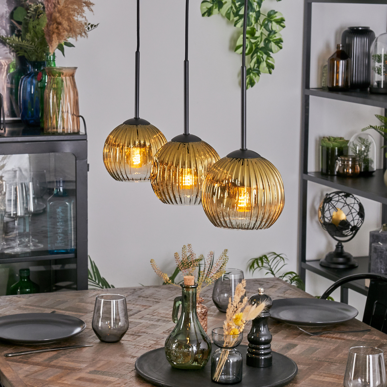 Drei goldene Pendelleuchten über einem Holztisch. Der Tisch ist mit Tellern, Gläsern und Dekorationsartikeln gedeckt. Regale im Hintergrund.