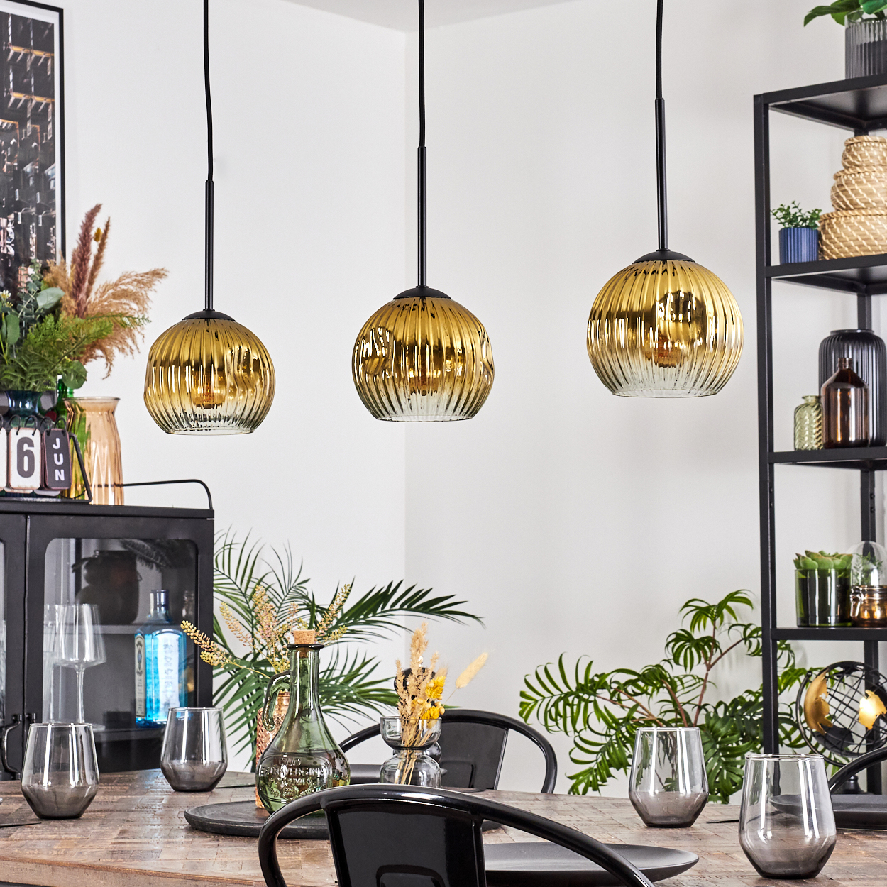 Esszimmer mit drei goldenen Pendelleuchten. Tisch mit Gläsern und Tellern gedeckt. Pflanzen und ein schwarzes Regal im Hintergrund.