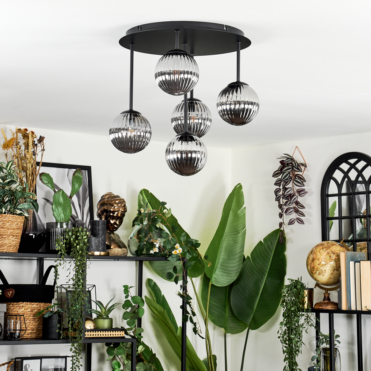 Ein modernes Wohnzimmer mit einem schwarzen Metallregal voller Pflanzen und Dekoration. Eine Hängelampe hängt von der Decke.