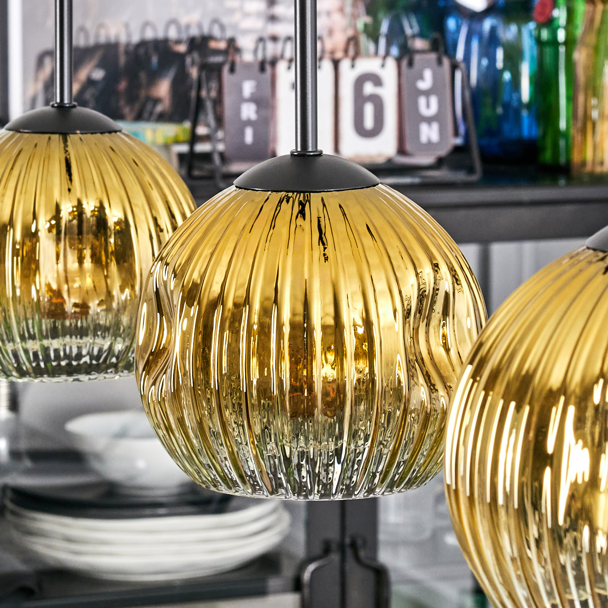 Drei goldene, gerippte Glas-Pendelleuchten hängen. Im Hintergrund sind Flaschen, ein Kalender und Teller.