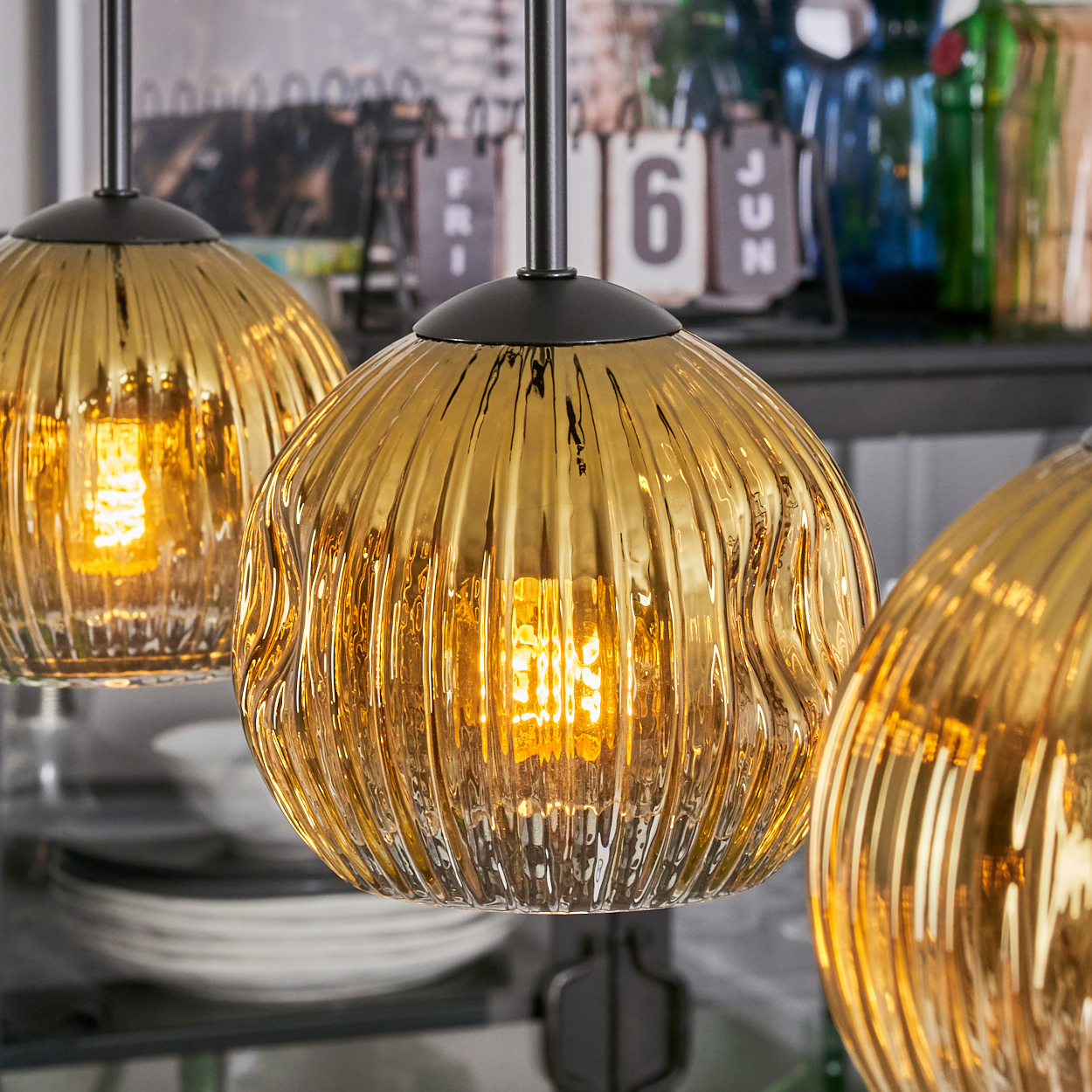 Drei goldene, gerippte Glas-Pendelleuchten mit freiliegenden Glühbirnen, die vor einem verschwommenen Hintergrund hängen.