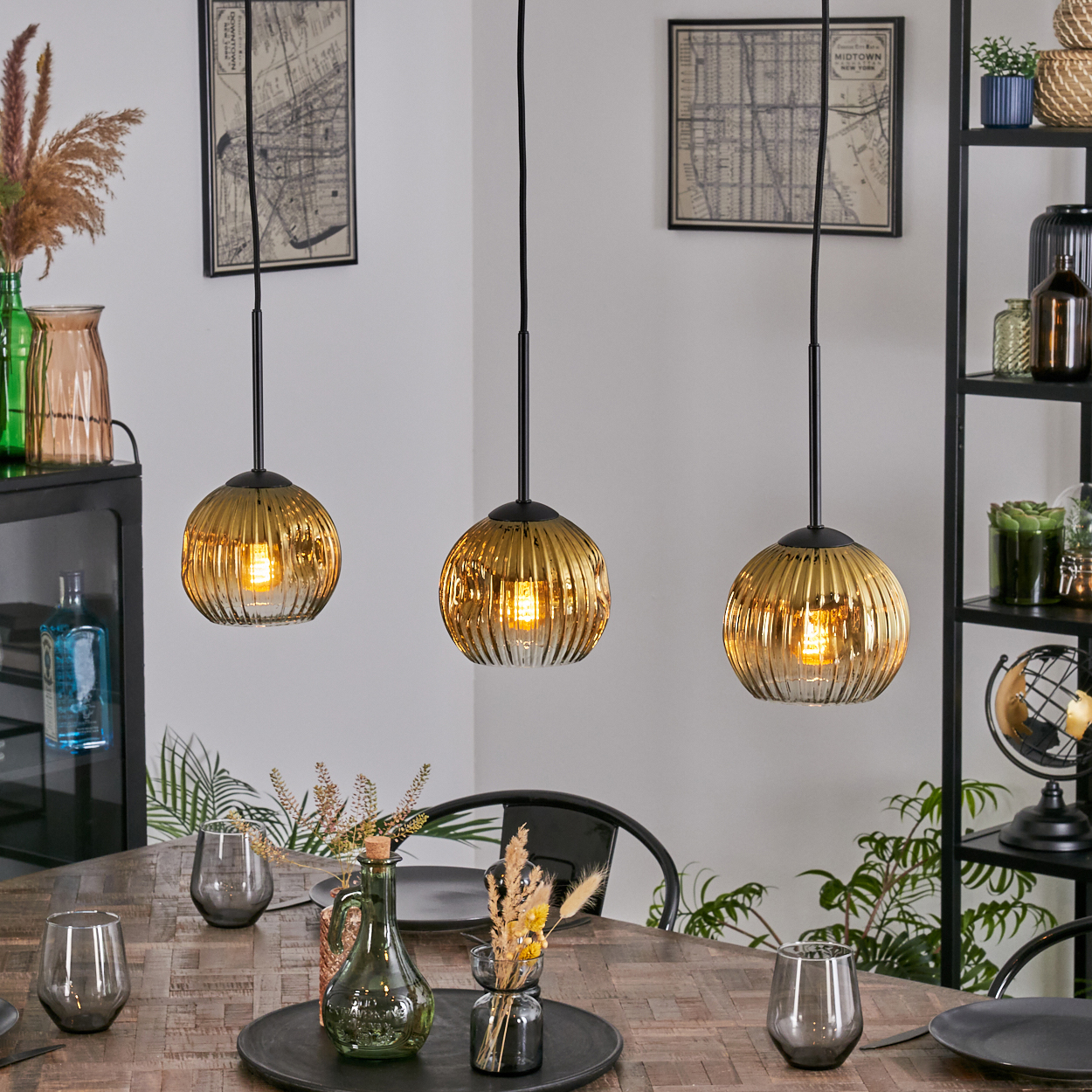Drei goldene Pendelleuchten hängen über einem Holztisch, der zum Abendessen gedeckt ist. Vasen und Gläser sind auf dem Tisch platziert.