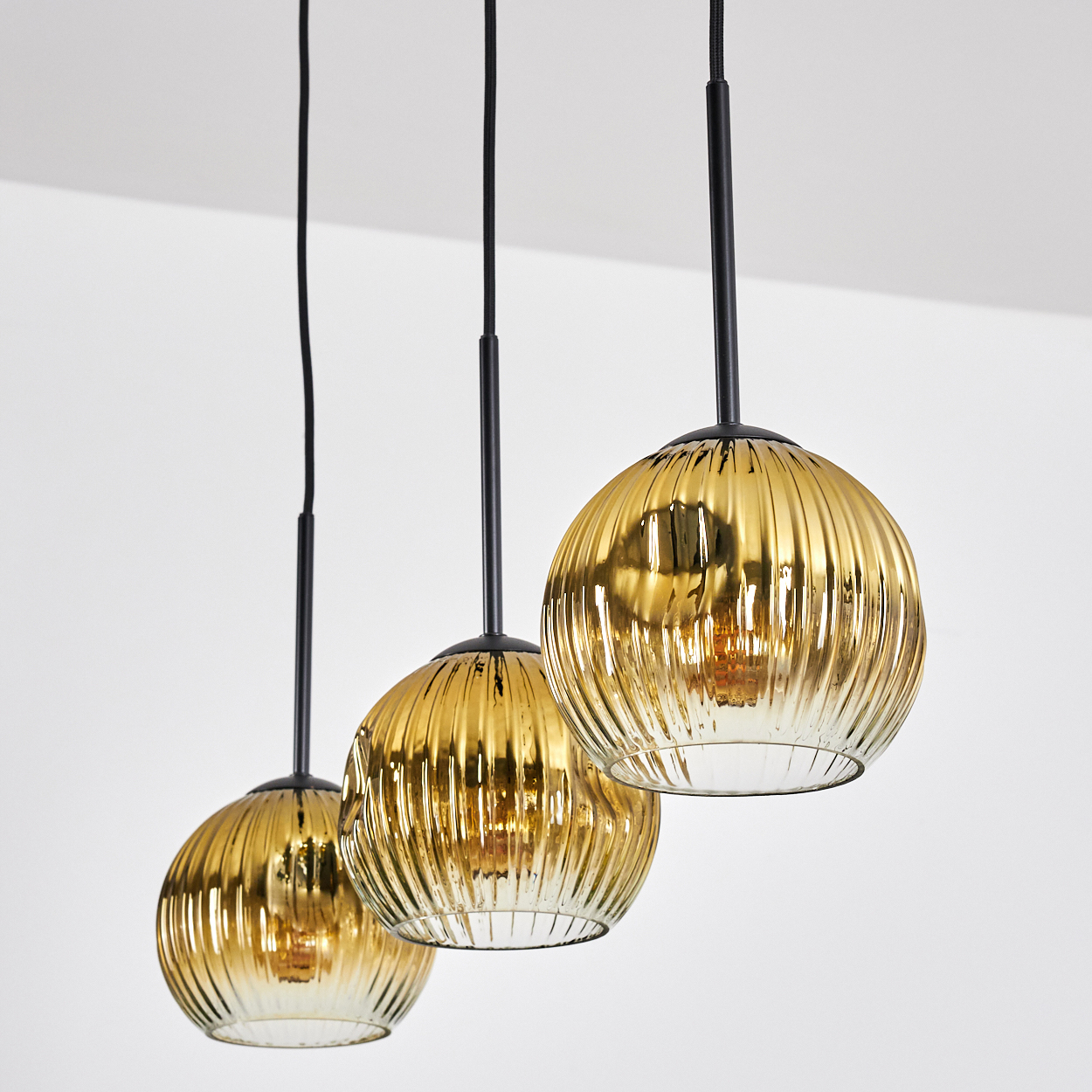 Drei goldene, geriffelte Glas-Pendelleuchten hängen vor weißem Hintergrund. Schwarze Kabel und Halterungen tragen die Leuchten.