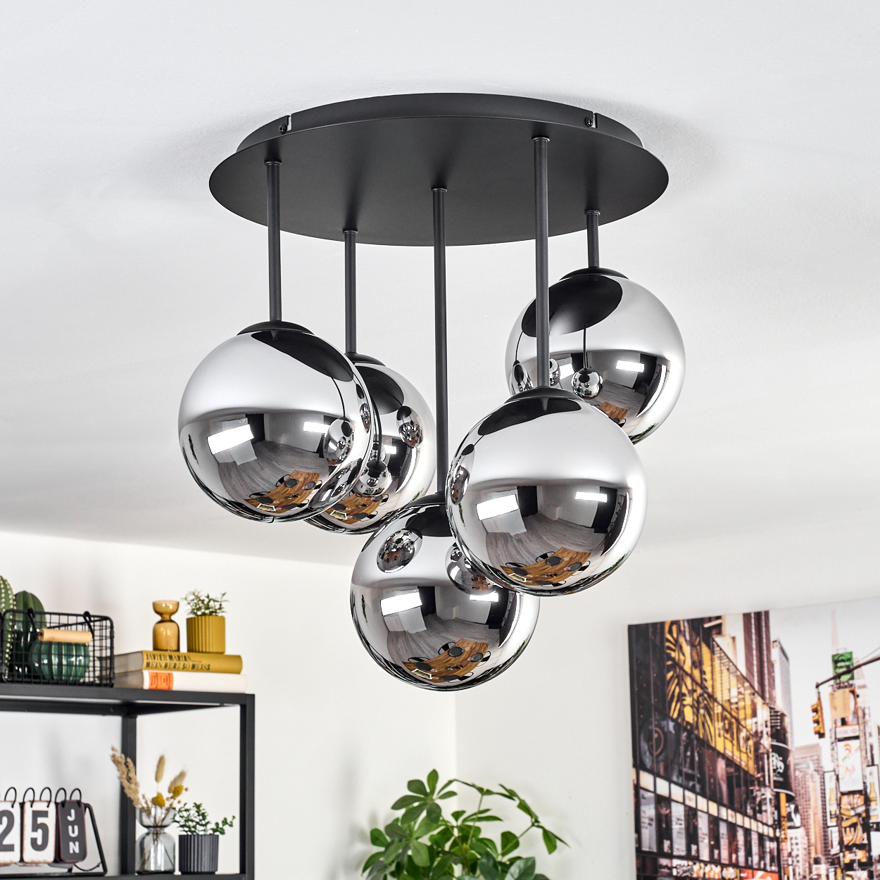 Eine moderne Deckenlampe mit fünf Chromkugeln. Die Kugeln reflektieren den Raum. Im Hintergrund ein Regal mit Dekorationen und ein Gemälde.