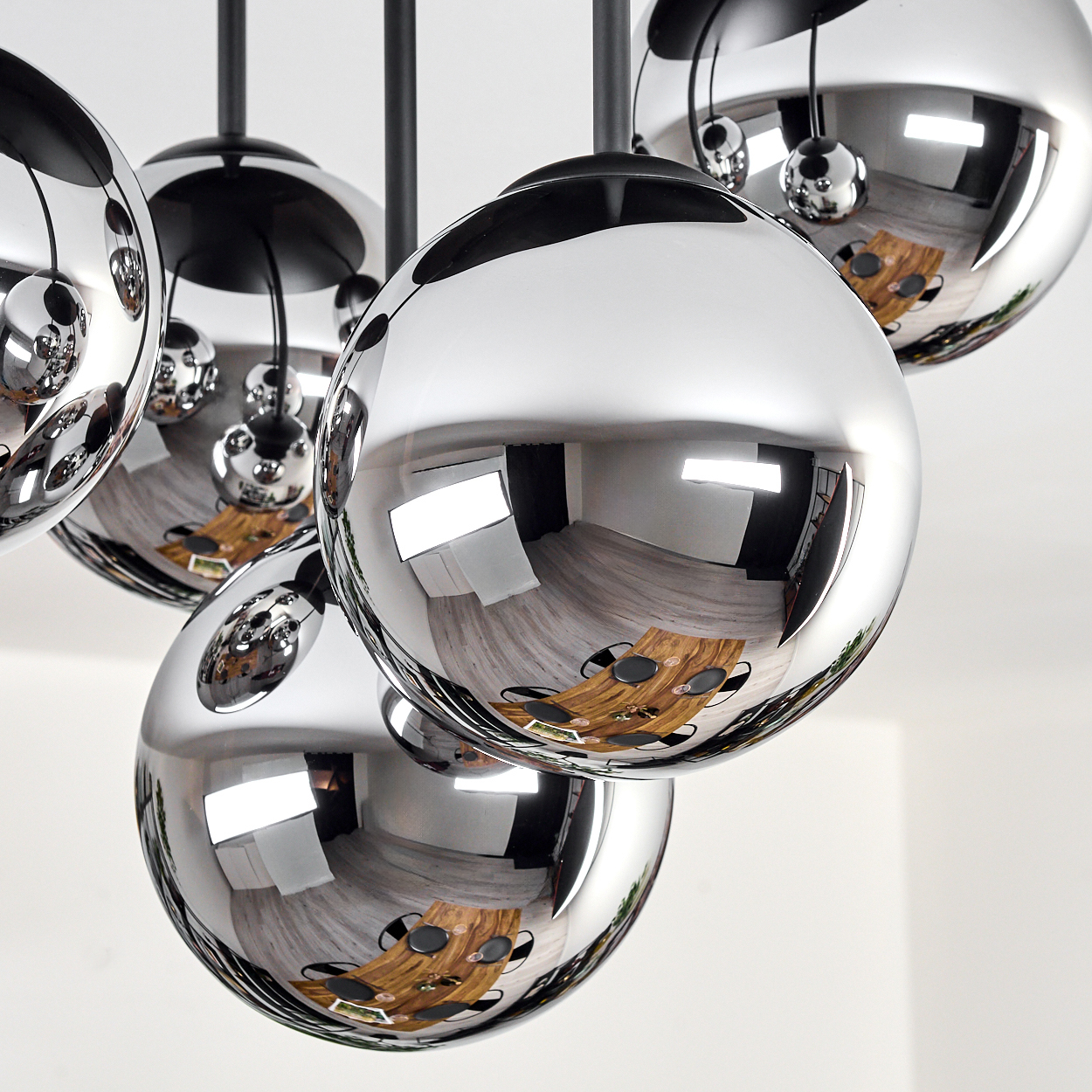 Chrom-Kugel-Kronleuchter, der einen Raum mit Tisch, Stühlen und Fenster reflektiert. Schwarze Stiele tragen die Kugeln. Weiße Decke.
