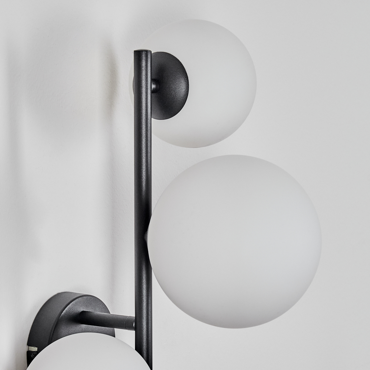 Eine moderne Wandlampe mit schwarzem Metallrahmen und drei kugelförmigen weißen Glasschirmen, an einer weißen Wand befestigt.