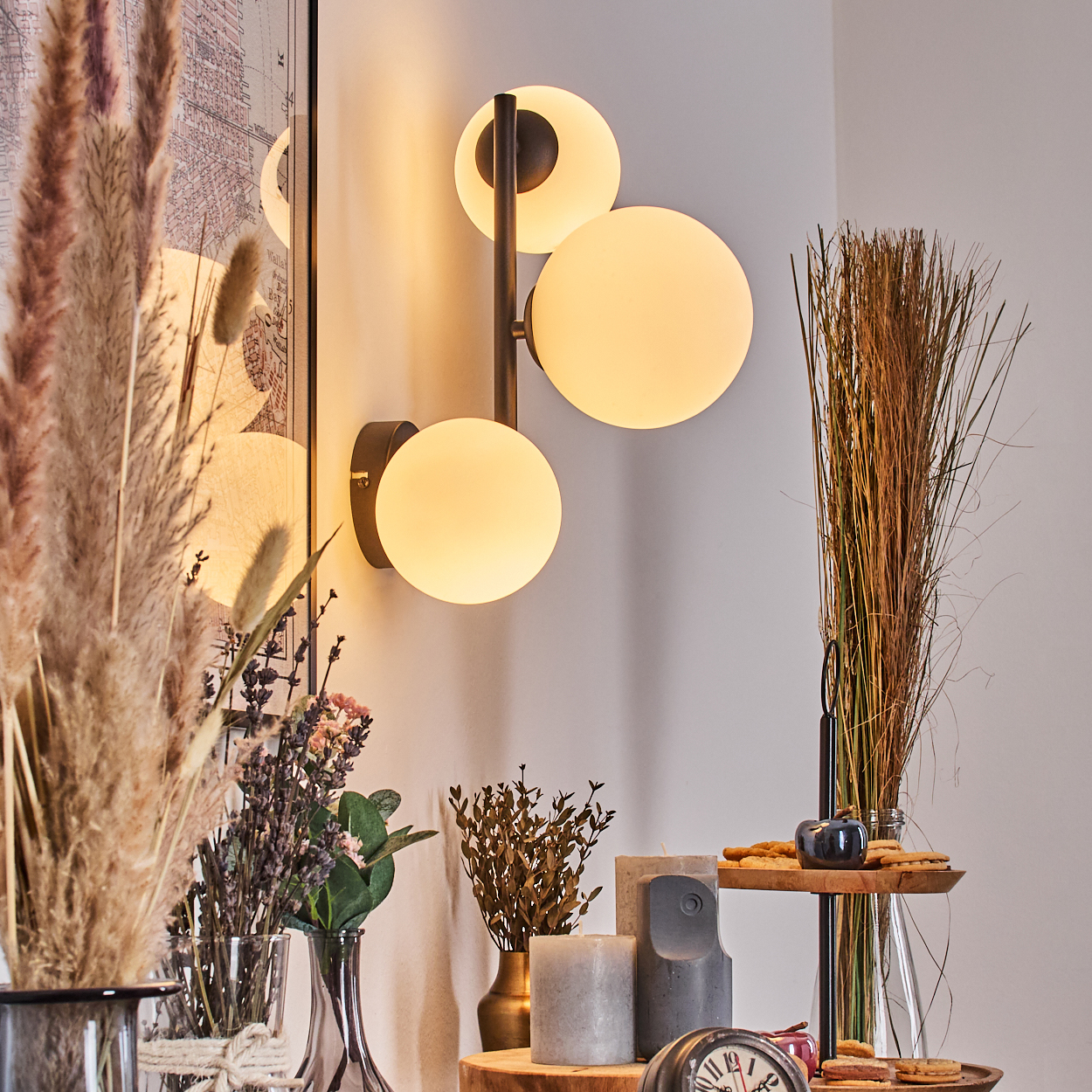 Dekorative Szene mit einer Wandlampe mit drei Glühbirnen, Trockenpflanzen, Kerzen und einem kleinen Holztablett mit Keksen und einem kleinen Kürbis.