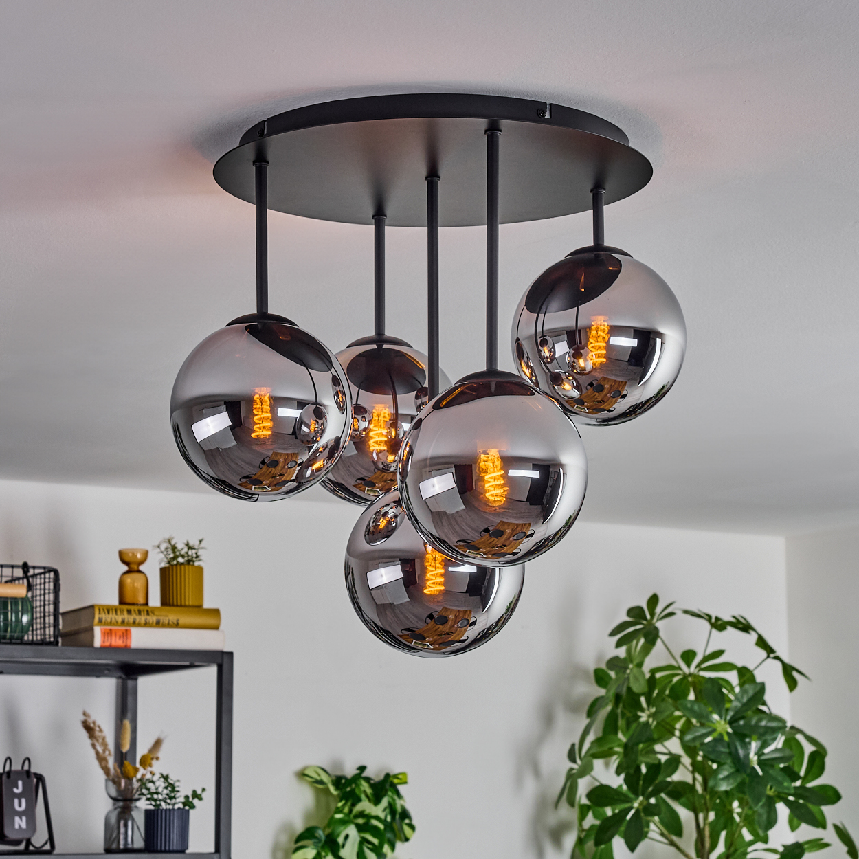 Eine moderne Deckenleuchte mit fünf kugelförmigen, rauchfarbenen Glasschirmen und sichtbaren Glühbirnen. Darunter ein Regal mit Dekoration.