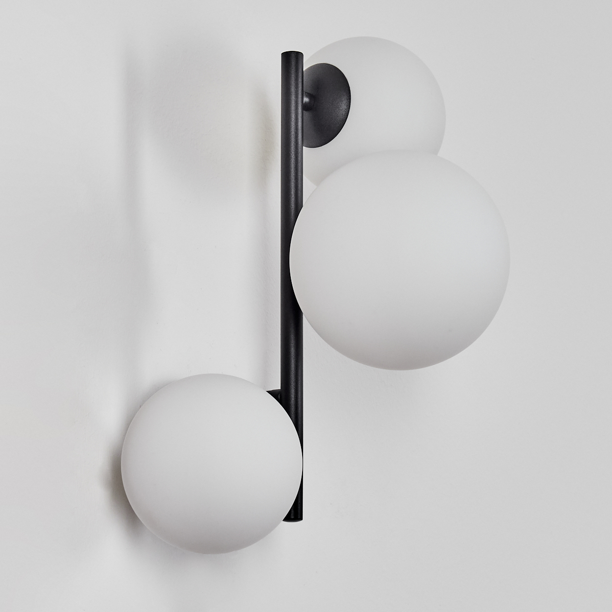 Eine moderne Wandlampe mit drei weißen, kugelförmigen Schirmen. Sie hat eine schwarze vertikale Halterung und einen schwarzen kreisförmigen Sockel.
