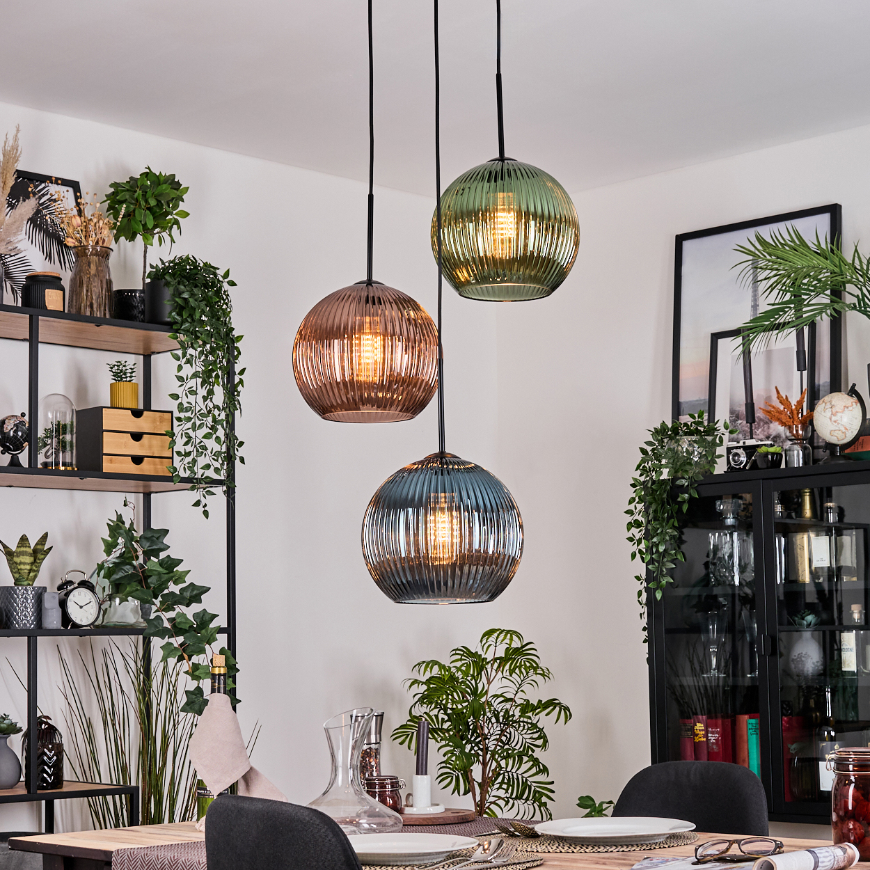 Drei Pendelleuchten mit Glasschirmen hängen über einem Esstisch. Das Zimmer hat ein Regal mit Pflanzen und Dekorationsartikeln.