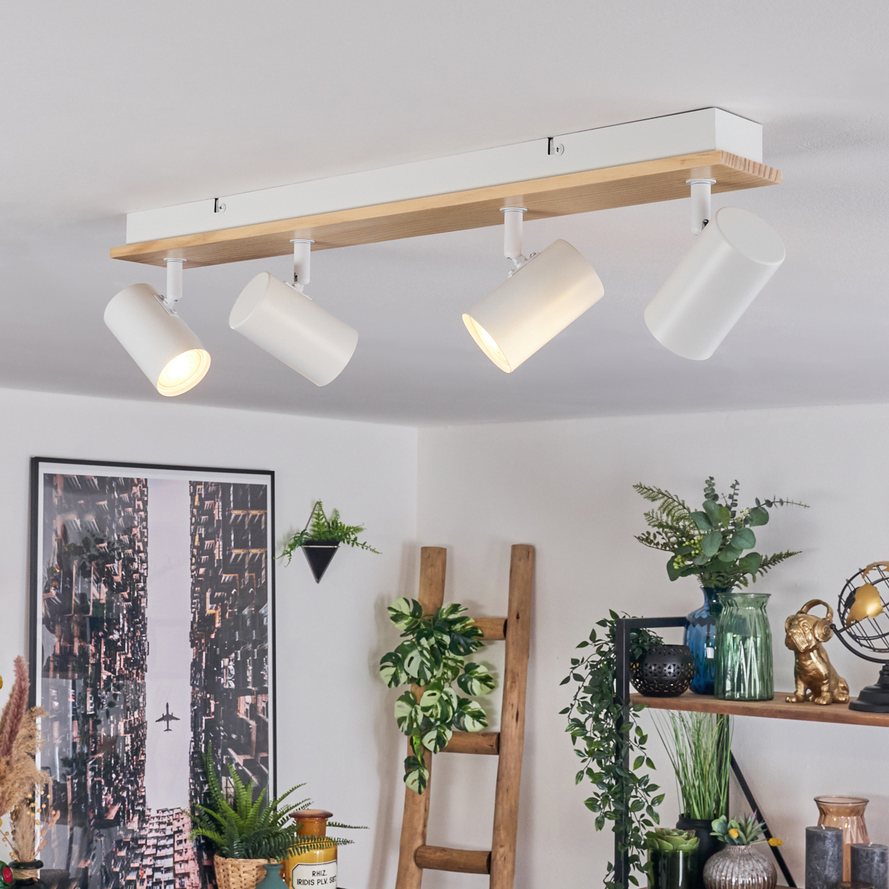 Eine weiße Deckenlampe aus Holz mit vier Strahlern ist an der Decke montiert. Im Raum befinden sich verschiedene Pflanzen und Dekorationsgegenstände.