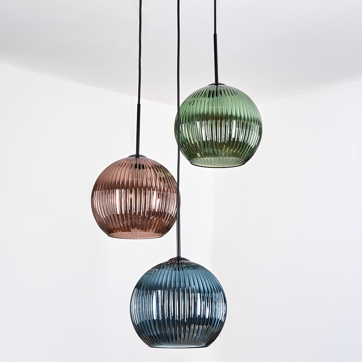 Drei kugelförmige Pendelleuchten hängen vor einer weißen Wand. Farben sind Kupfer, Grün und Blau. Die Leuchten sind gerippt und haben schwarze Kabel.