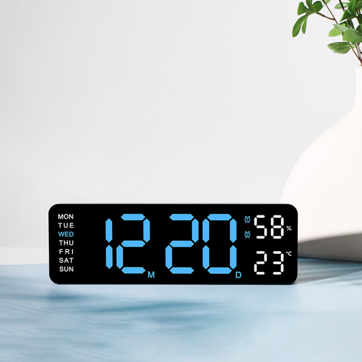 Eine Digitaluhr zeigt 12:20, 58 % Luftfeuchtigkeit und 23 °C an. Die Uhr steht auf einer Oberfläche mit Vase und Pflanze im Hintergrund.