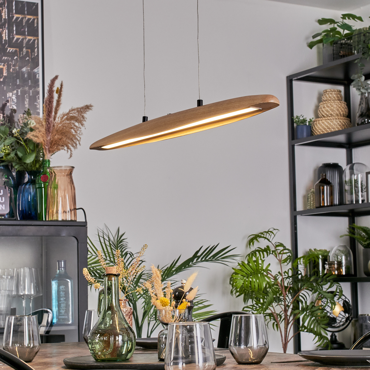 Ein modernes Esszimmer mit einer Holzlampe, Pflanzen und Gläsern auf dem Tisch. Regale befinden sich im Hintergrund.